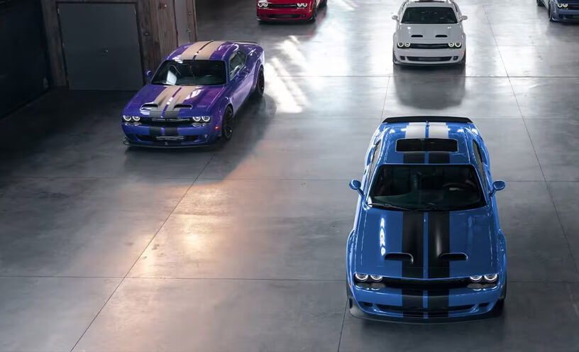 Six 2023 Dodge Challenger models parked in a commercial garage. They include a purple Challenger SRT Hellcat, a blue Challenger SRT Hellcat with black center stripes, a red Challenger SRT Hellcat, a white Challenger SRT Hellcat and more.